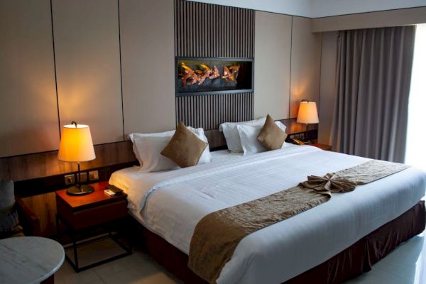 A modern hotel room features a king-size bed, two bedside tables with lamps, wall art above the headboard, and a small table by the window.