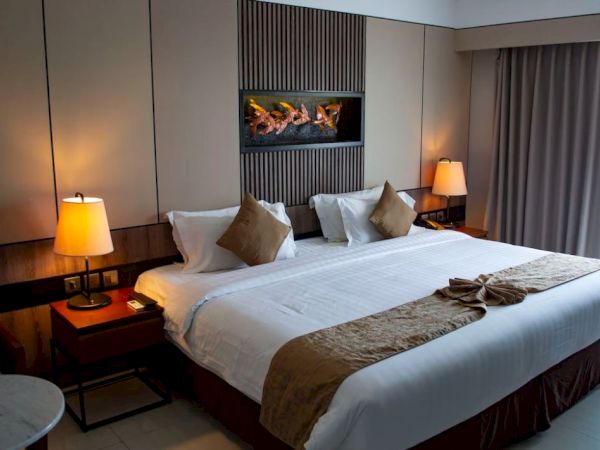 A modern hotel room features a king-size bed, two bedside tables with lamps, wall art above the headboard, and a small table by the window.