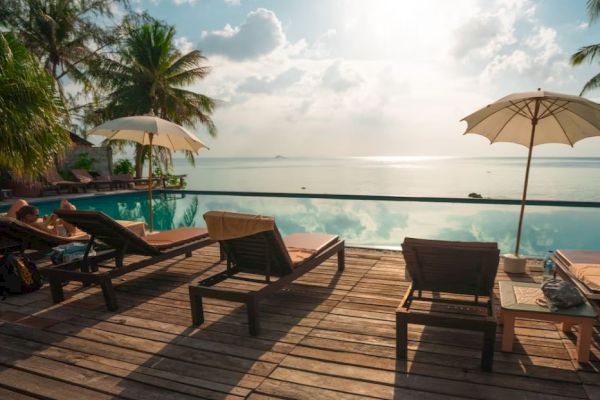 A serene poolside scene with lounge chairs, umbrellas, and an infinity pool overlooking a calm sea, surrounded by palm trees, under a bright sky.