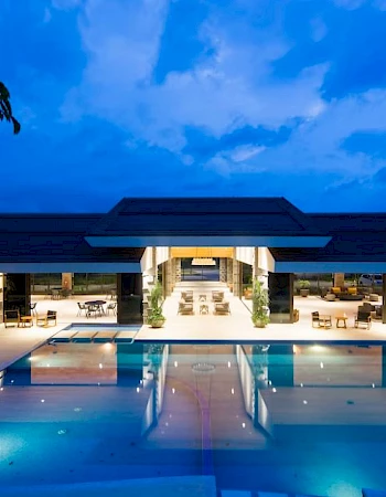 An illuminated modern building with an expansive swimming pool in the foreground, set against a backdrop of a twilight sky.