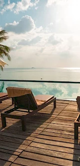 A serene poolside scene with lounge chairs and umbrellas overlooking a calm ocean under a partly cloudy sky, surrounded by tropical plants.