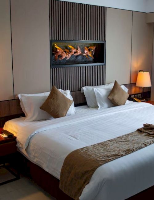A hotel room with a large bed, two bedside tables with lamps, decorative pillows, a wall-mounted fireplace, and curtains covering a window.
