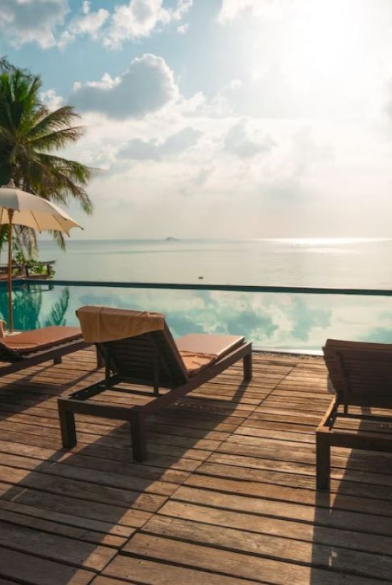 A serene poolside scene with lounge chairs, umbrellas, palm trees, and an infinity pool overlooking a calm ocean under a bright, partly cloudy sky.