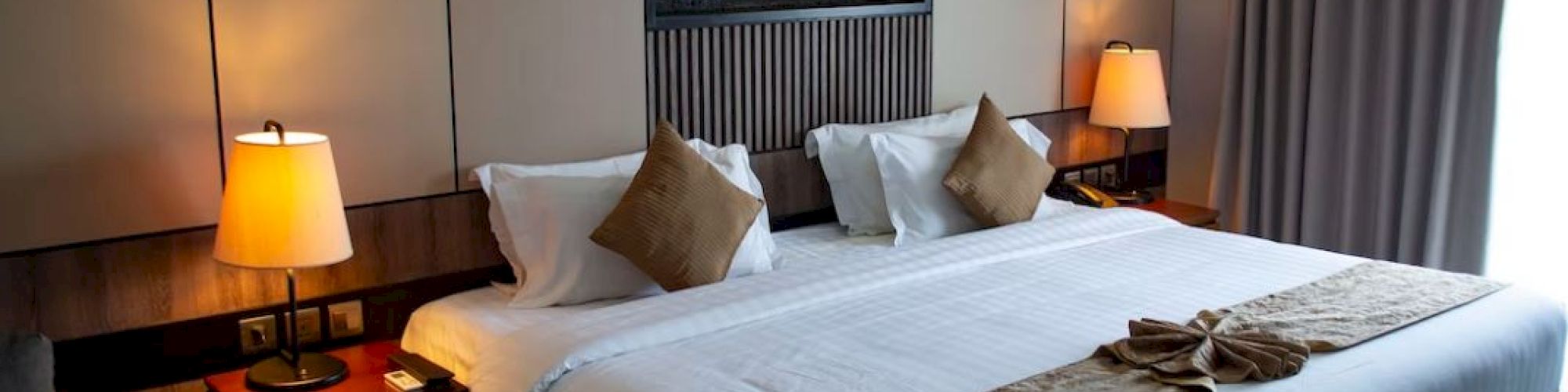 A well-lit hotel room features a large bed with white linens and beige accents, bedside tables with lamps, and a cozy ambiance.