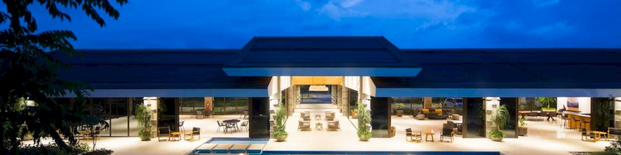 An illuminated modern building with an outdoor swimming pool at dusk, featuring lounge chairs and a spacious open area.