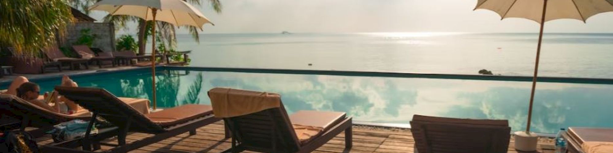 A serene poolside scene with lounge chairs and umbrellas, overlooking a calm body of water. There's a wooden deck and tropical trees around.