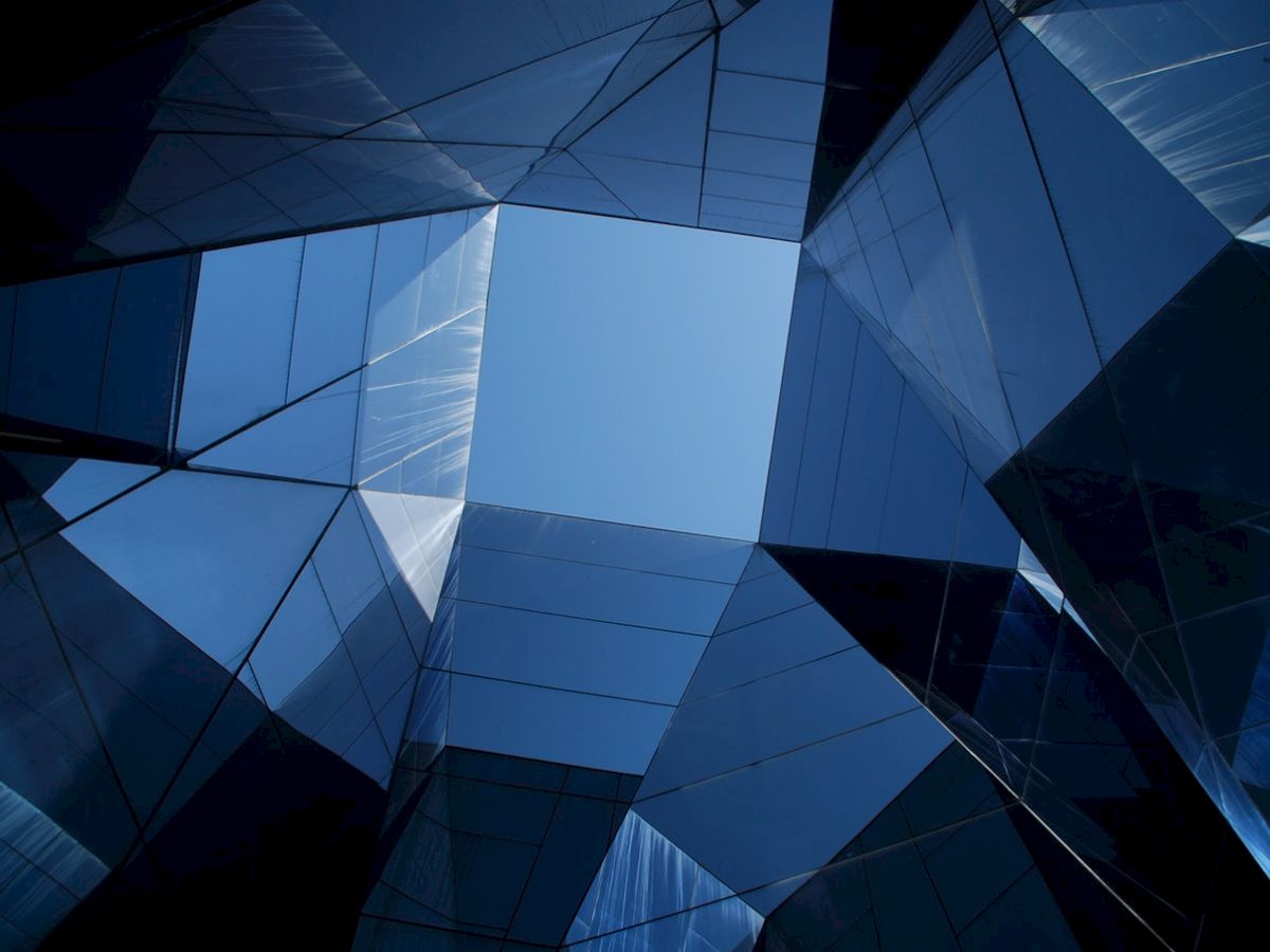 The image shows modern architectural structures made of reflective glass panels, with a view of the sky through a geometrical opening.