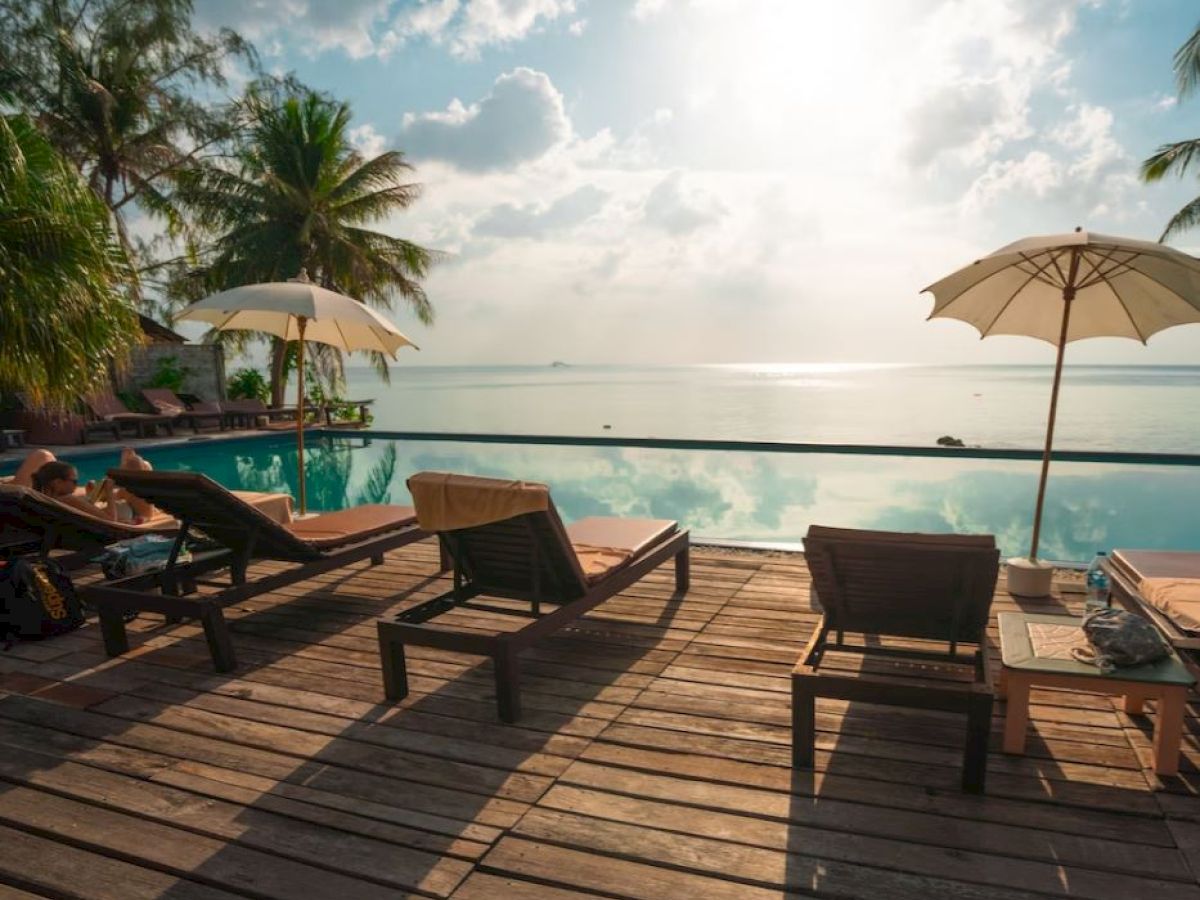 This image shows a serene poolside with sun loungers, umbrellas, and palm trees overlooking a calm sea under a partly cloudy sky, creating a relaxing ambiance.