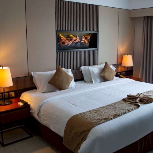 A cozy hotel room features a large bed with white linens and brown pillows, two bedside tables with lamps, and a small fireplace above the bed.