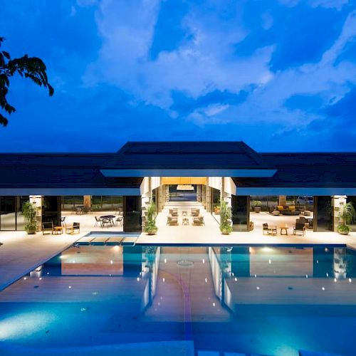 A luxurious outdoor pool area at night, with adjacent seating and dining areas under a covered pavilion, surrounded by trees.