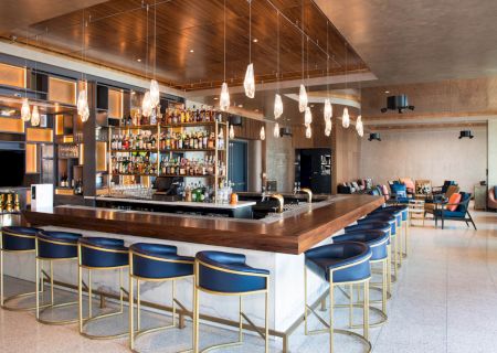 A modern bar with a large counter, blue stools, a well-stocked shelf of alcohol, contemporary lighting, and a comfortable seating area.