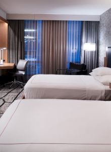 A modern hotel room with two neatly made beds, a wall-mounted TV, a desk with a chair, and stylish decor featuring patterned wallpaper and curtains.