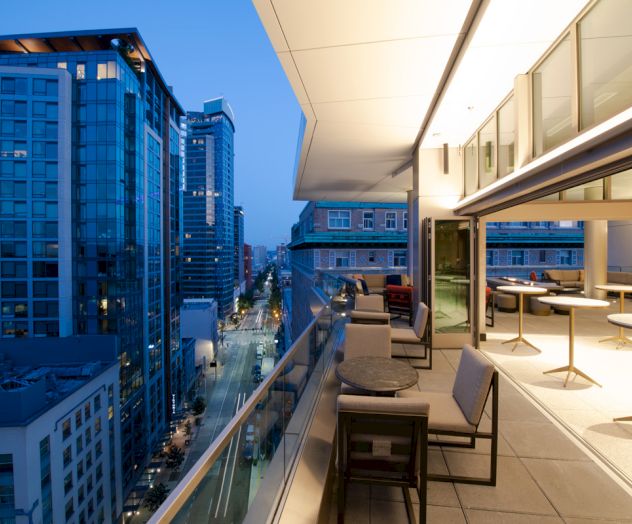 This image shows a modern balcony with seating and tables overlooking a cityscape with tall buildings and a well-lit street in the evening.