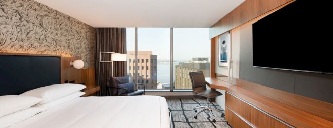 A modern hotel room with a large bed, flat-screen TV, desk, and window showing a cityscape outside. The decor features wood accents and neutral tones.