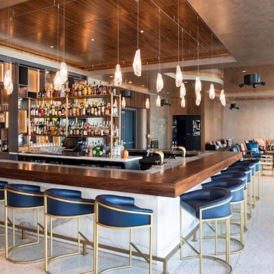 A stylish bar with modern seating, a variety of drinks behind the counter, pendant lighting, and additional lounge seating in the background.