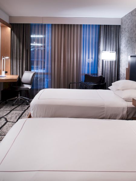 A modern hotel room with two neatly made beds, a wall-mounted TV, a desk with a chair, bedside lamps, and floor-to-ceiling curtains.