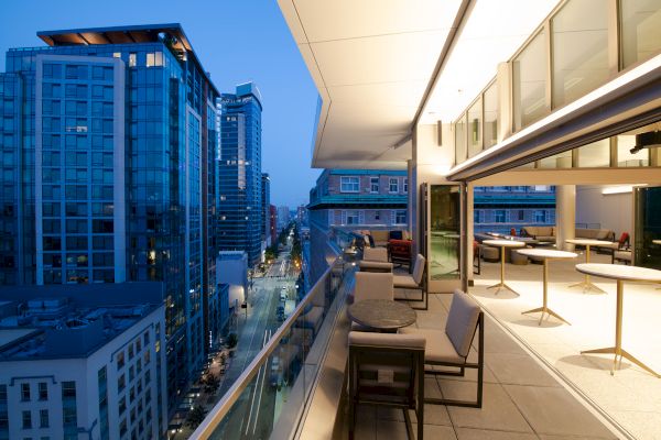 A modern city rooftop terrace at dusk, featuring outdoor seating and tables with tall buildings and a street visible in the background.