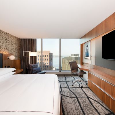 A modern hotel room with a large bed, wall-mounted TV, desk, chair, and a window with a city view.