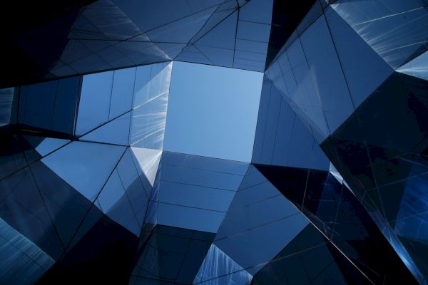 The image shows a geometric architectural structure composed of glass panels, reflecting the sky and creating an abstract visual effect.