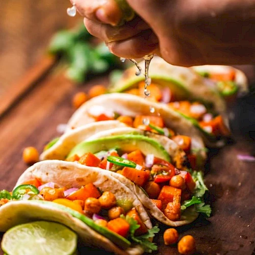 The image shows a person squeezing lime juice over a row of tacos filled with chickpeas, vegetables, and garnished with avocado slices and cilantro.
