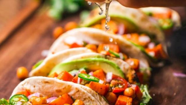 The image shows a person squeezing lime juice over a row of tacos filled with chickpeas, vegetables, and garnished with avocado slices and cilantro.