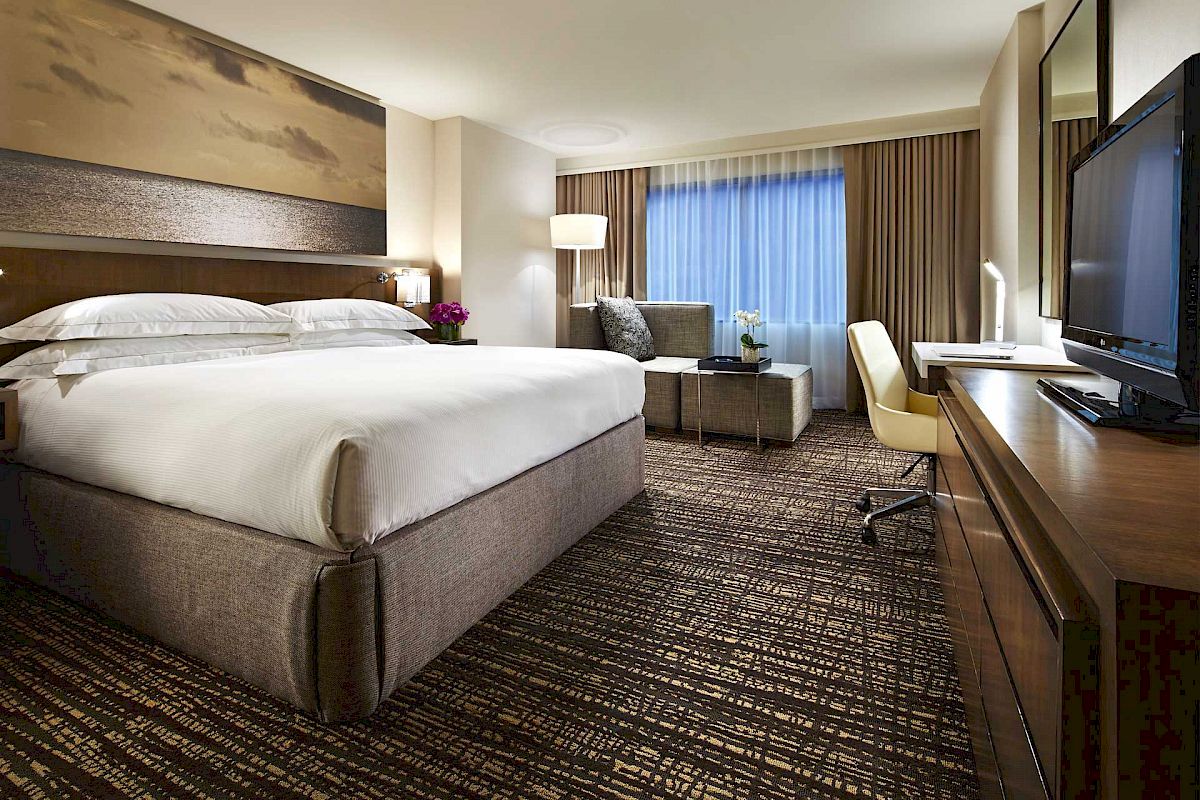 This image shows a modern hotel room with a large bed, a desk, a chair, a sofa, and a TV. The window has curtains partially drawn.