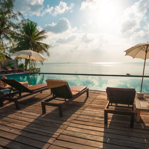 Sun loungers and umbrellas on a wooden deck by an infinity pool overlook a serene ocean with a bright sky and palm trees in the background.