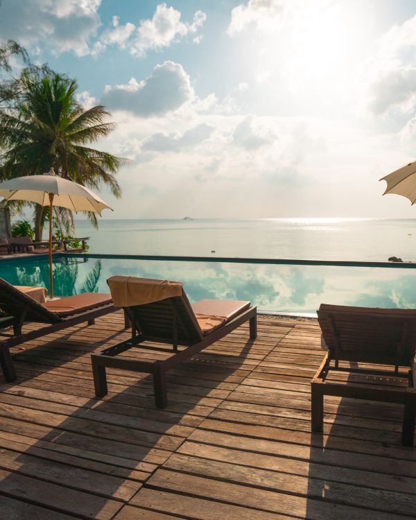 Sun loungers and umbrellas on a wooden deck by an infinity pool overlook a serene ocean with a bright sky and palm trees in the background.