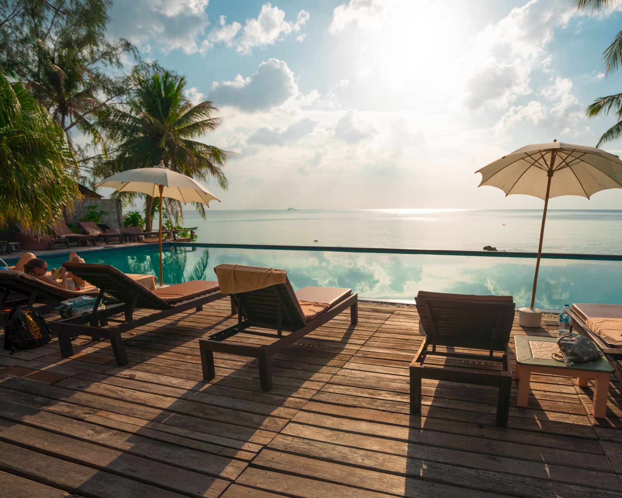 The image shows a tranquil poolside scene with lounge chairs, umbrellas, and a stunning view of the ocean under a partly cloudy sky.