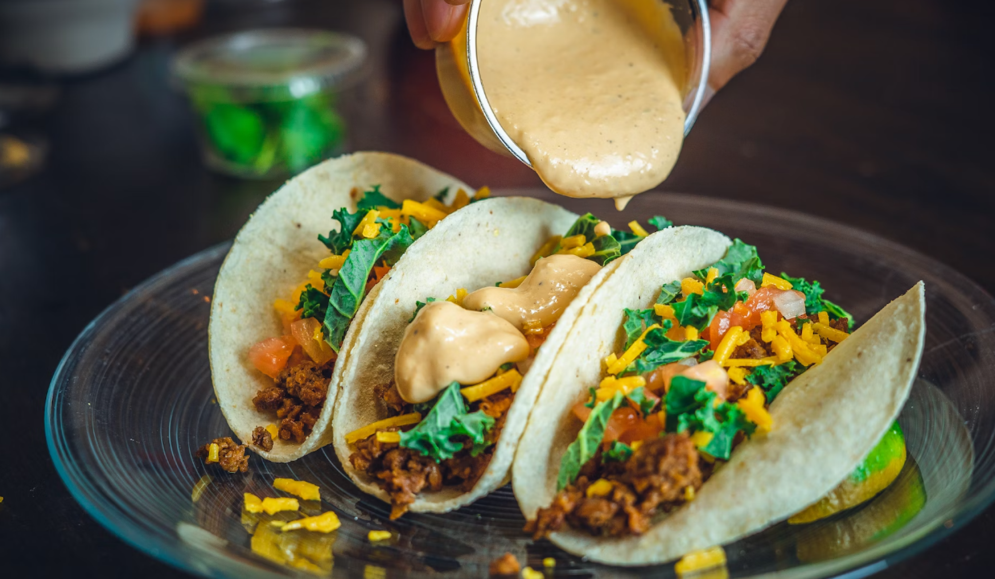 The image shows three tacos filled with ground meat, shredded cheese, and vegetables, with a hand pouring a creamy sauce over them.