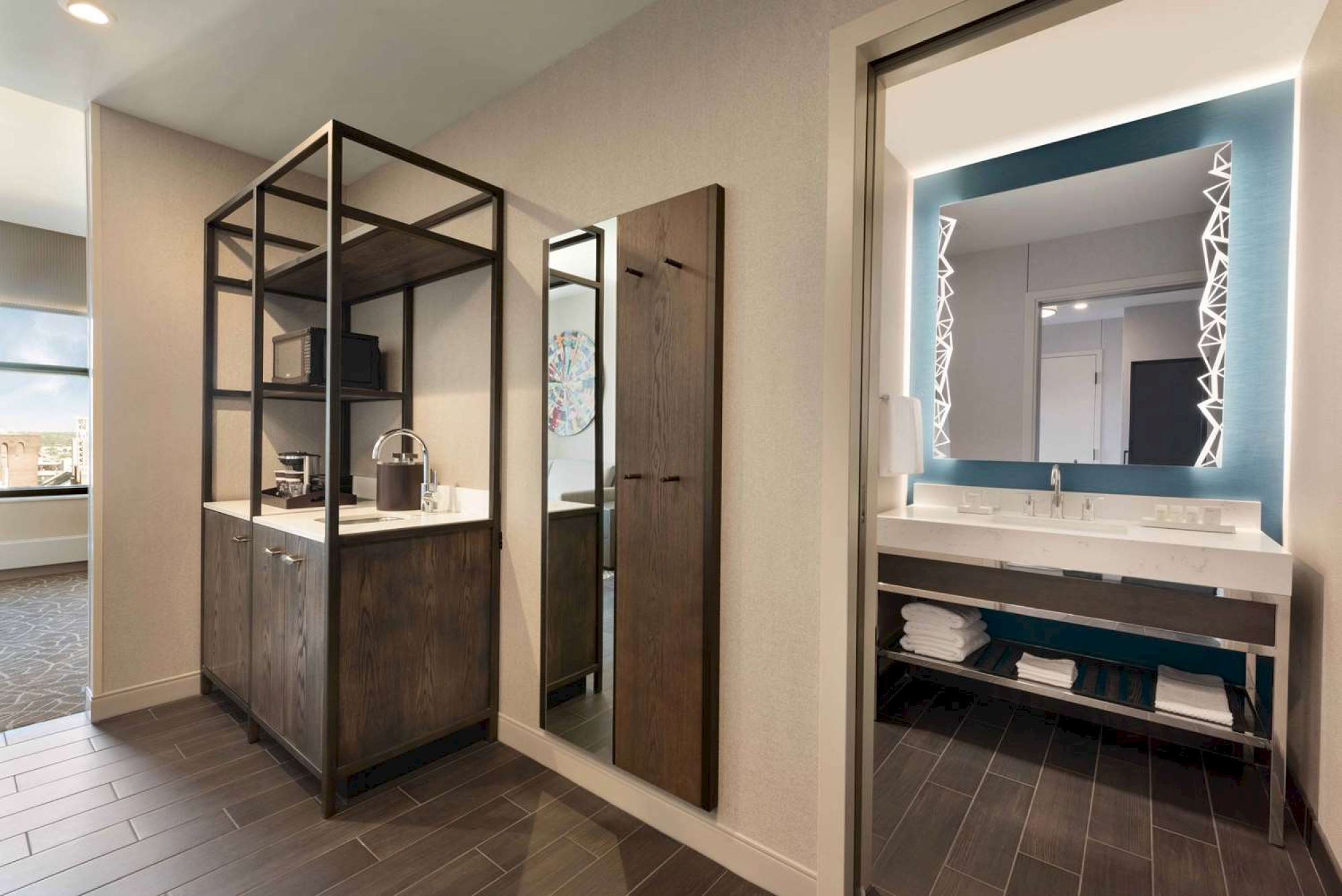 The image shows a modern hotel room corner with a kitchenette and a bathroom. The kitchenette has a coffee machine, cabinets, and a mirror.