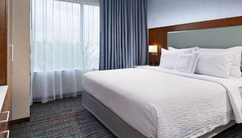 A modern hotel room with a large bed, white bedding, a window with blue and white curtains, bedside lamps, and carpeted flooring.