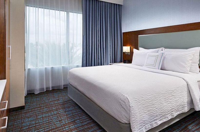 A modern hotel room with a large bed, white bedding, a window with blue and white curtains, bedside lamps, and carpeted flooring.