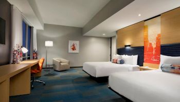 The image shows a modern hotel room with two beds, a desk, a chair, a large window, and a colorful abstract painting on the wall.