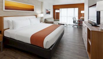 This image shows a modern hotel room with a king-sized bed, a sofa, a desk, a TV, and a large window with curtains.