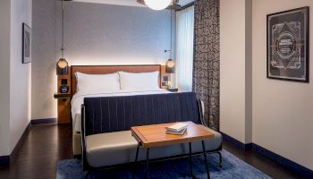 A modern hotel room with a large bed, a small sofa, a table with a book, stylish lighting, artwork on the walls, and a blue rug.