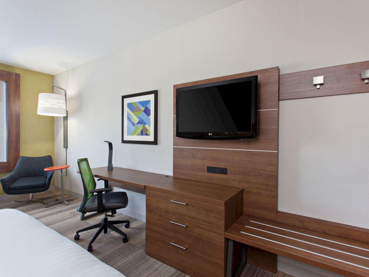 Modern hotel room featuring a desk with a green office chair, wall-mounted TV, chair with ottoman, and floor lamp. Abstract art on the wall.