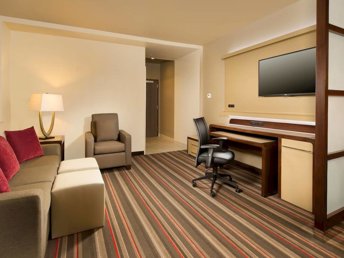 The image shows a modern hotel room with a sofa, armchair, desk, office chair, lamp, and wall-mounted TV. The room has striped carpet flooring.