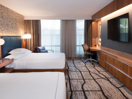 The image shows a modern hotel room with two beds, a TV, a desk with a chair, a patterned carpet, and large windows with sheer curtains.