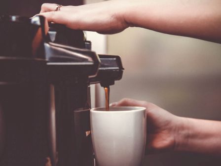 A close-up shot of hands making coffee with an espresso machine. One hand is operating the machine while the other is holding a white mug.