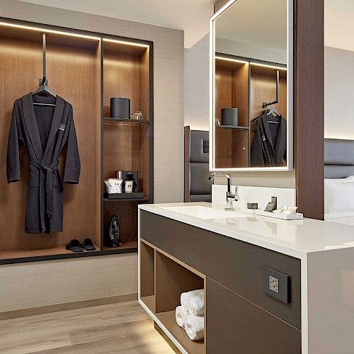 A modern hotel room features a neatly organized open closet, a sink area with a large mirror, and a bed with a padded headboard.