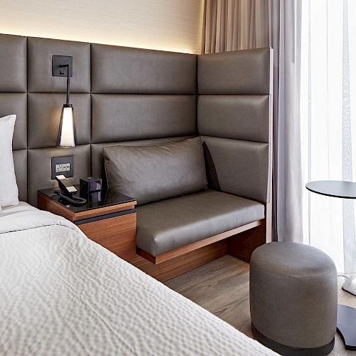 A modern hotel room corner with a bed, cushioned bench, armchair, side table, and a window letting in natural light.