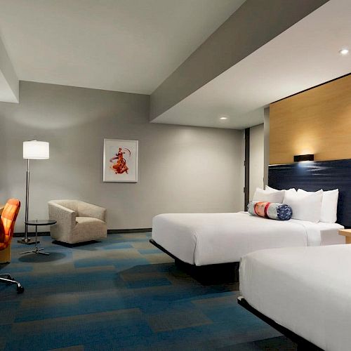 This image shows a modern hotel room with two double beds, a desk with an orange chair, a beige armchair, and a floor lamp.