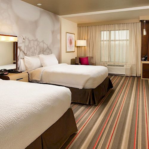 This image shows a hotel room with two beds, a desk, a chair, a TV, and a striped carpet. The room has modern decor and a window with curtains.