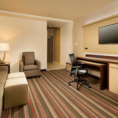 The image shows a modern hotel room with a sofa, armchair, ottoman, lamp, desk, office chair, and a wall-mounted TV.