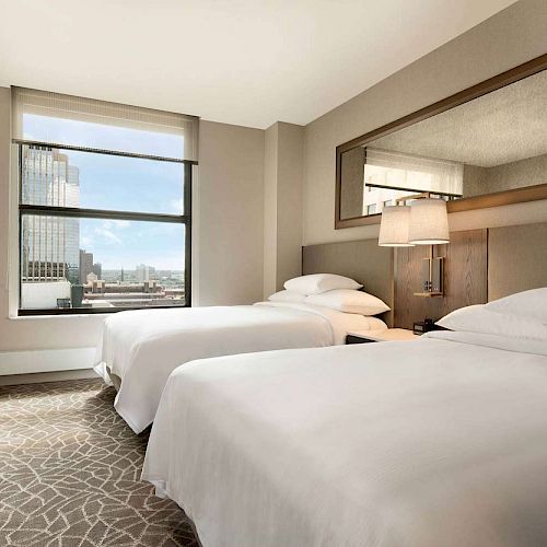 The image shows a modern hotel room with two double beds, a window with a city view, a wall-mounted mirror, and a desk with a chair.