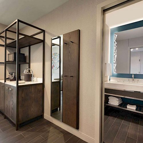 The image shows a modern hotel room with a kitchenette area and a sleek bathroom featuring a large illuminated mirror and double sink vanity.