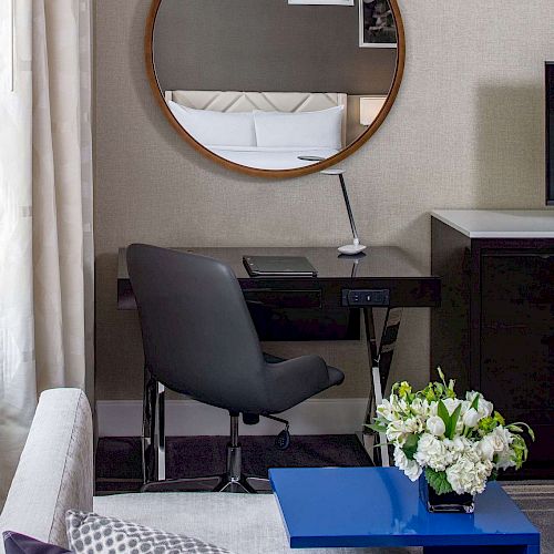 The image shows a modern, cozy workspace with a round mirror, a black desk, a grey chair, and a blue side table adorned with fresh flowers.