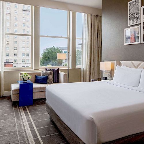 This image shows a modern hotel room with a large bed, desk area, seating near the window, and minimalist decor featuring framed art.