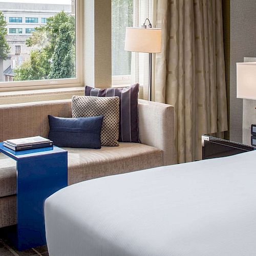 A cozy hotel room with a bed, a sofa by the window, navy blue pillows, a bright blue side table with books, and a view of trees outside.